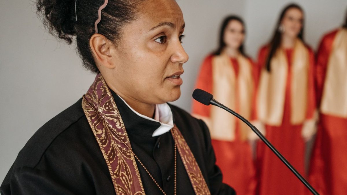 Pastora fala em uma igreja diante de um coral, durante uma atividade religiosa.