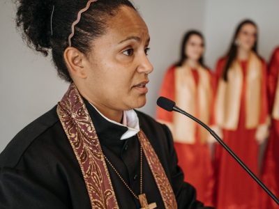 Pastora fala em uma igreja diante de um coral, durante uma atividade religiosa.