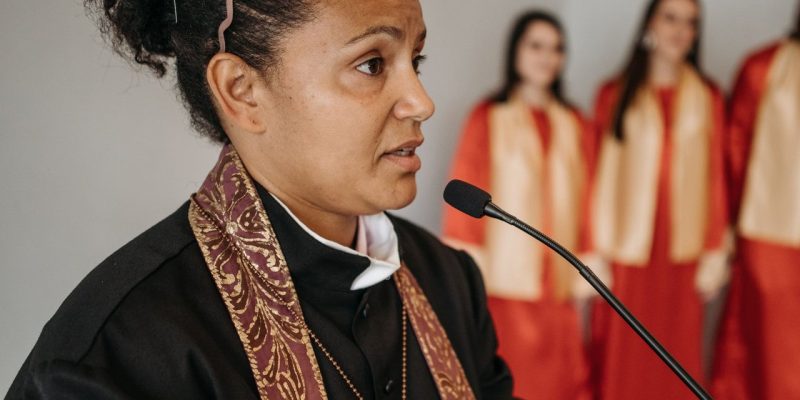 Pastora fala em uma igreja diante de um coral, durante uma atividade religiosa.