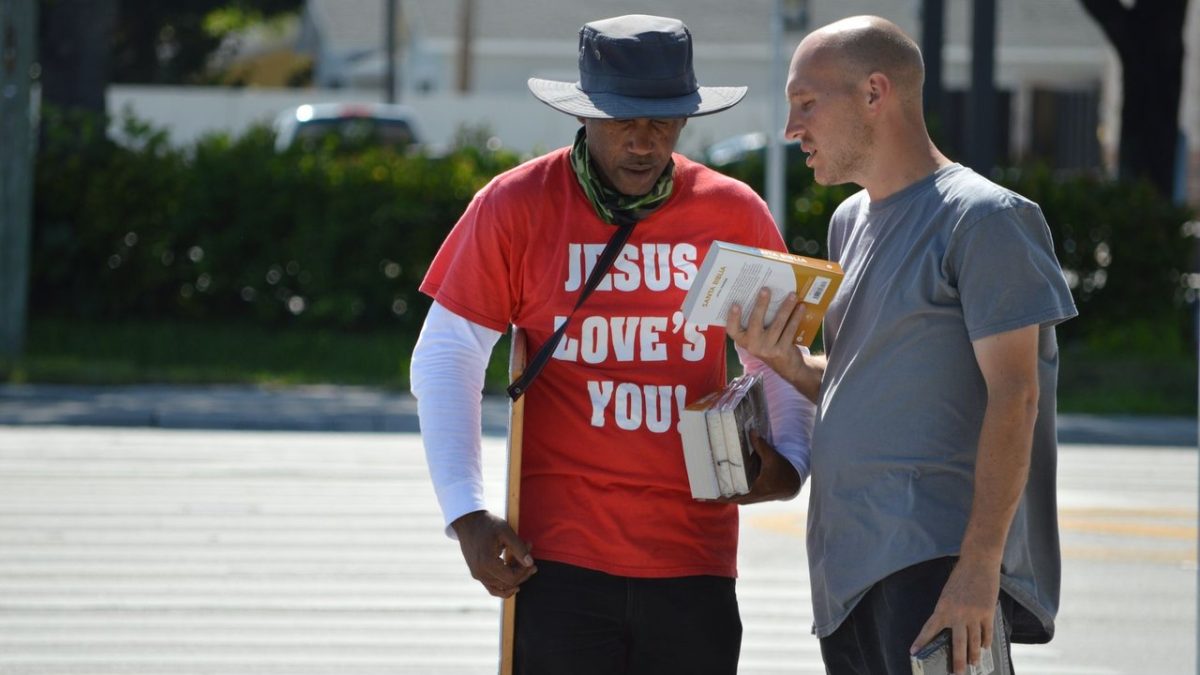 Dois homens conversam em uma rua, segurando livros durante uma abordagem religiosa