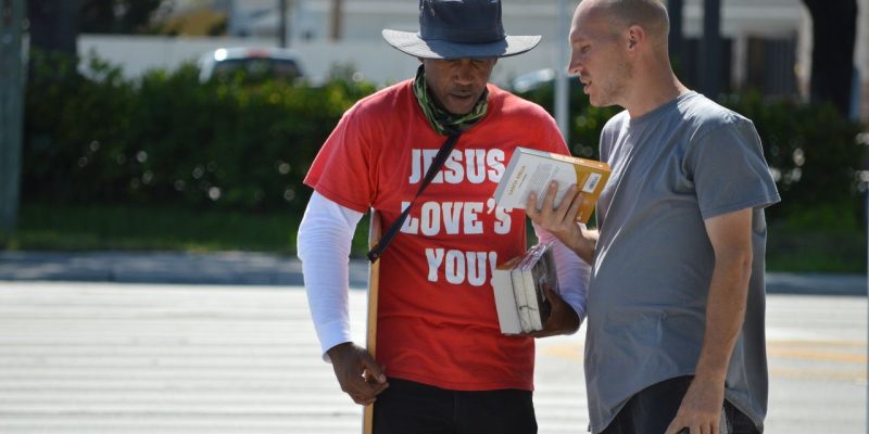 Dois homens conversam em uma rua, segurando livros durante uma abordagem religiosa